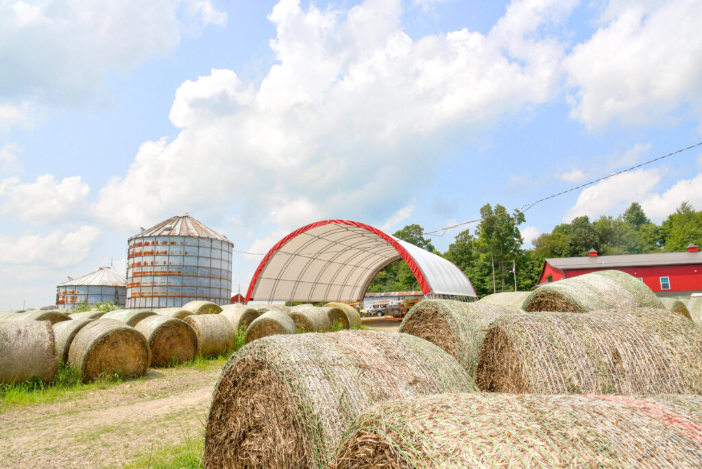 Round hay bales outside an open-front hoop building on a farm.
