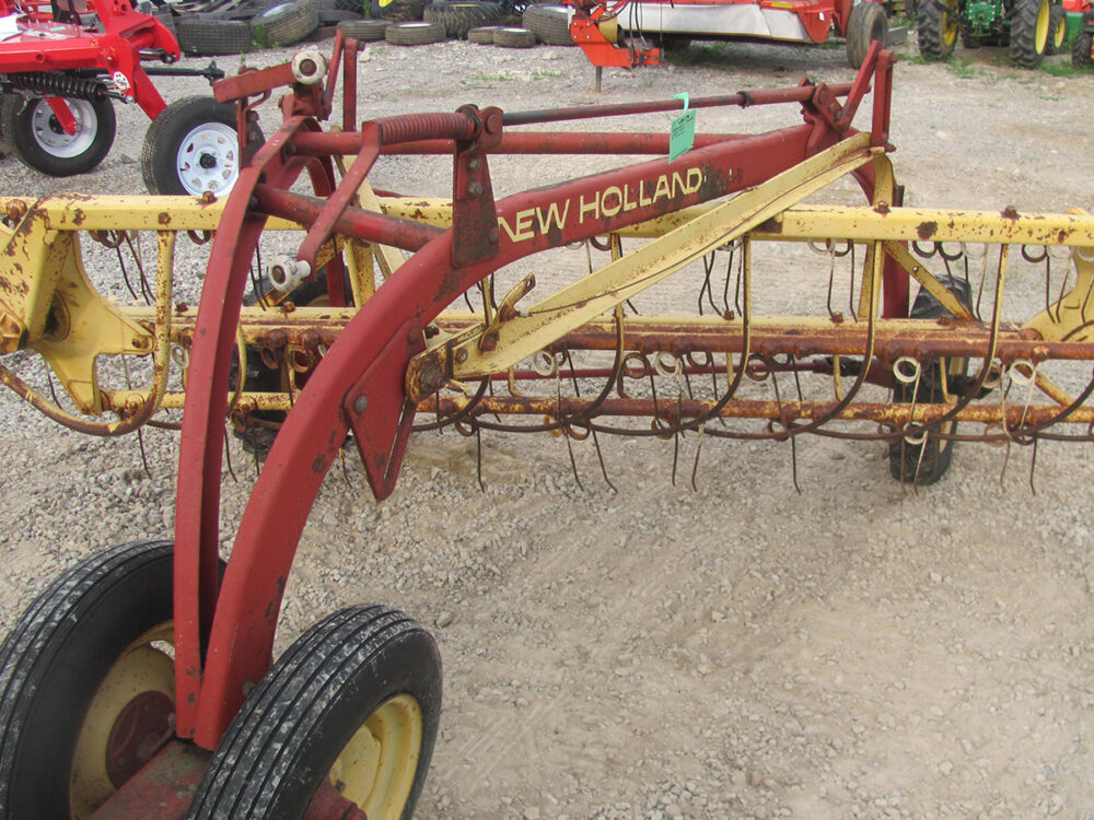 Used New Holland 256 Rollabar Rake with dolly wheels; one wheel needs replacement and some tines are missing.