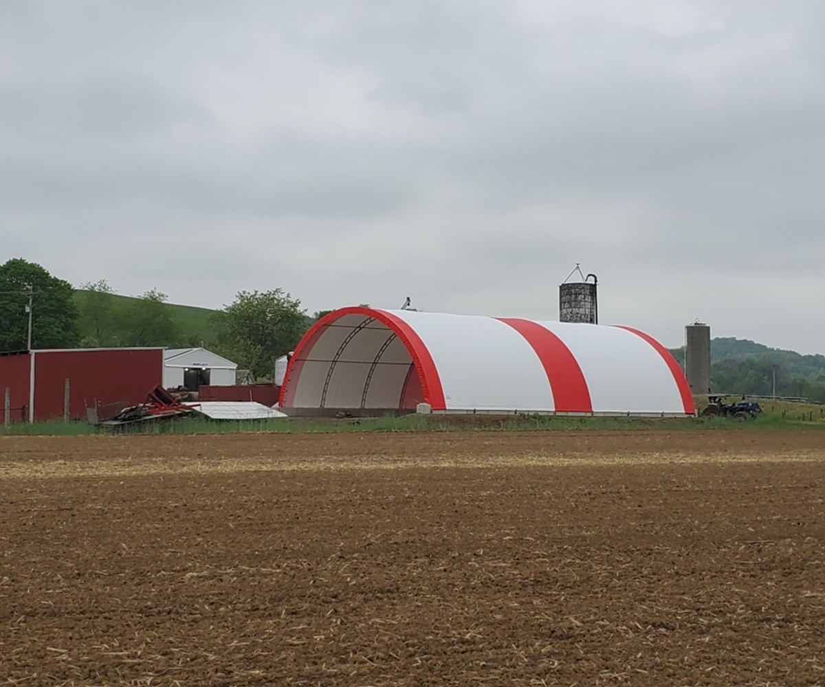 Red-and-white hoop building on farm field beside barns under cloudy sky.
