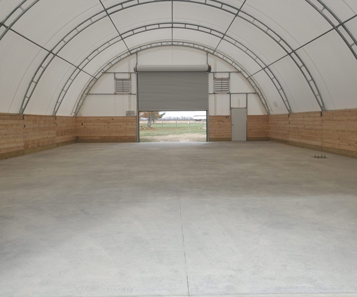 Inside hoop building with arched steel frame, concrete floor, and large roll-up door.