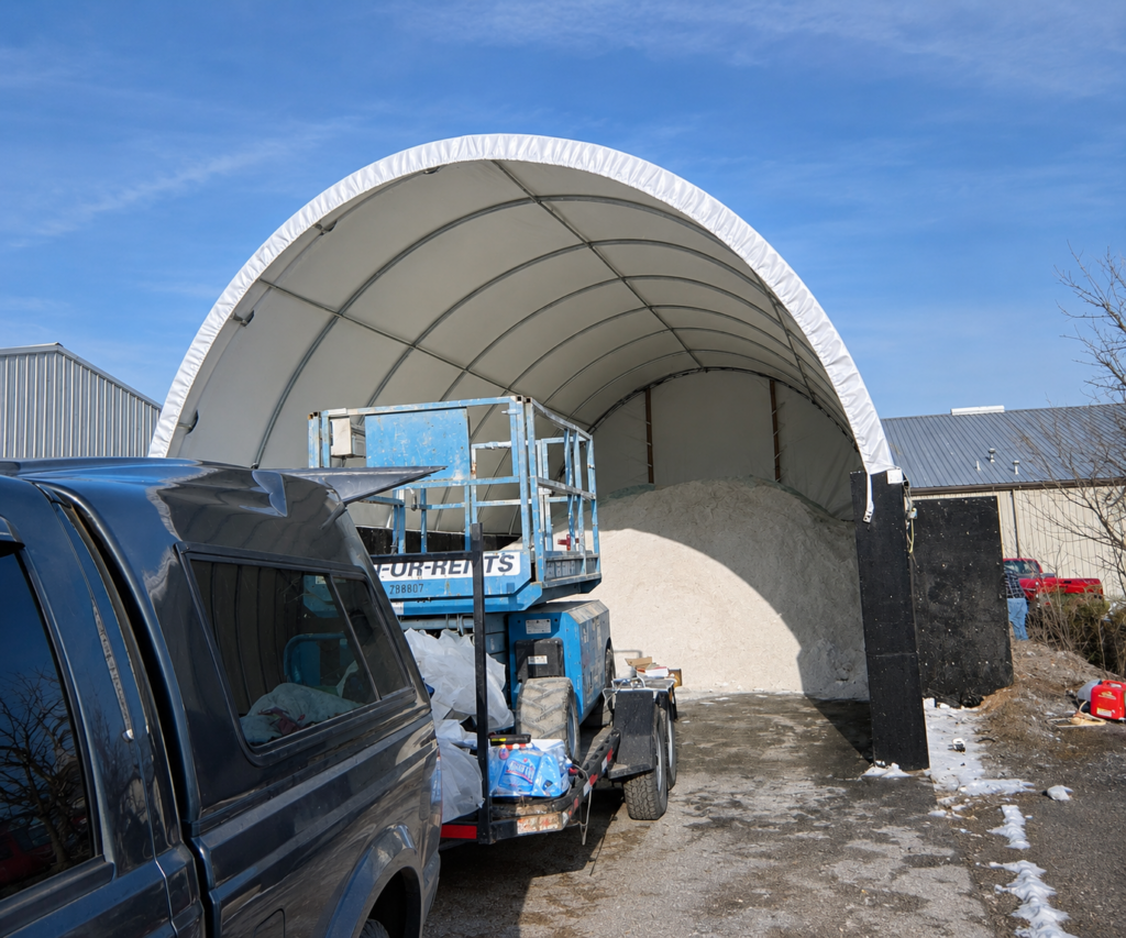 Open-front fabric storage building with a bulk material pile inside and equipment at the entrance.